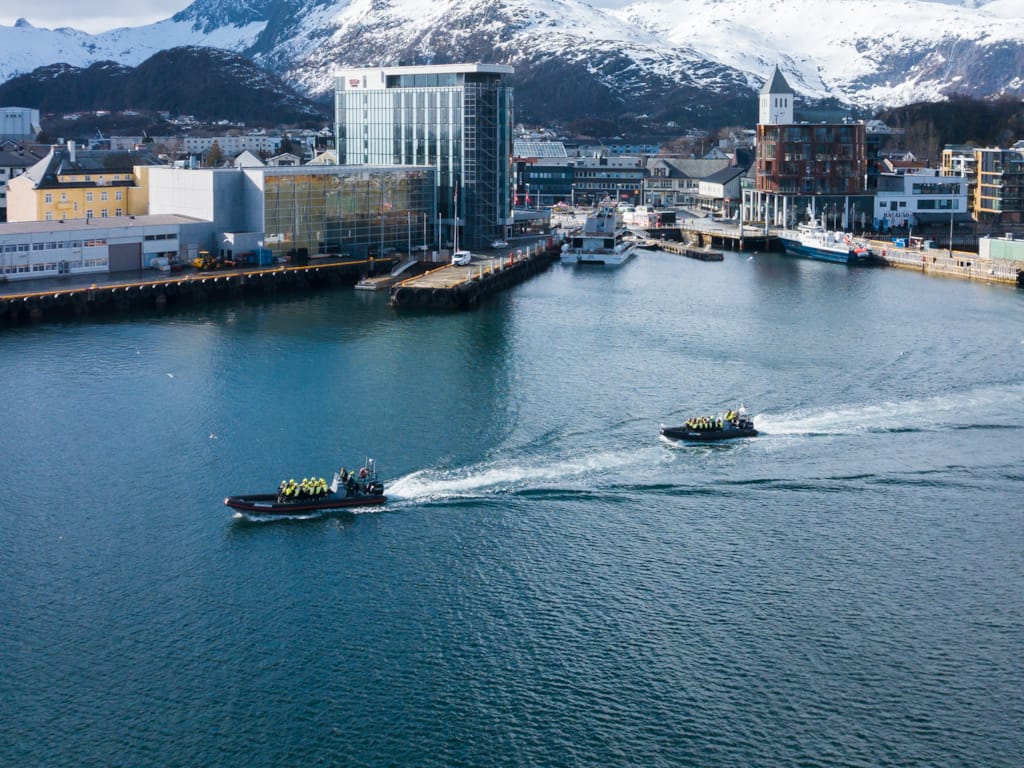 Zwei Rib-Boote auf dem Weg aus dem Hafen in Richtung Meer. Im Hintergrund sind das Thon Hotel Lofoten, das Zentrum von Svolvær und schneebedeckte Berge zu sehen.
