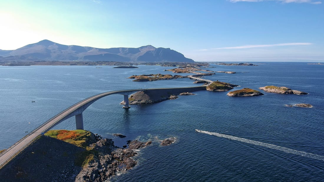 The Atlantic Road seen from above. A boat is driving on the water. Mountains can be seen in the background.