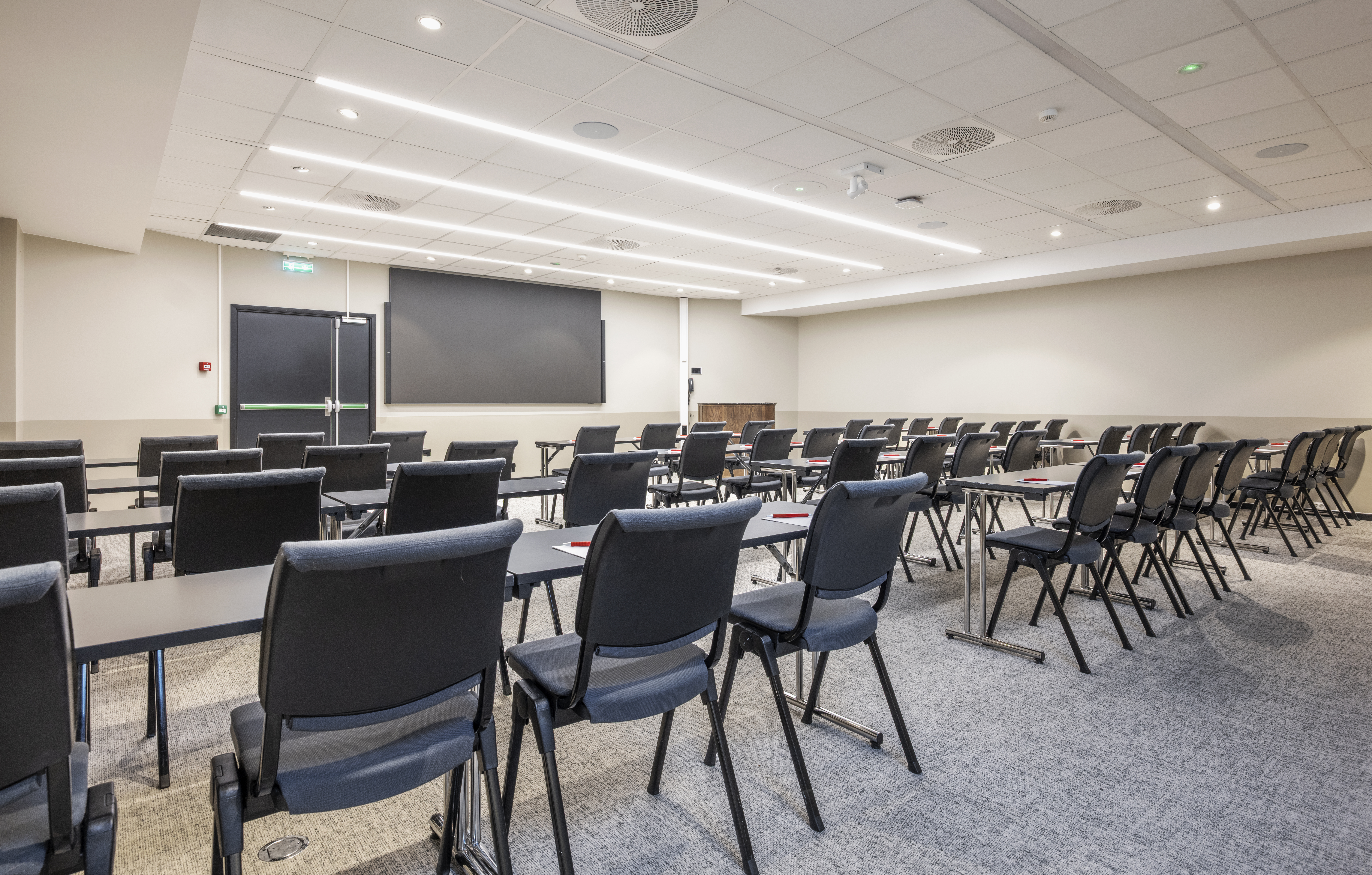 Konferanserom oppsett med stoler bak bord og en skjerm forrest i rommet.