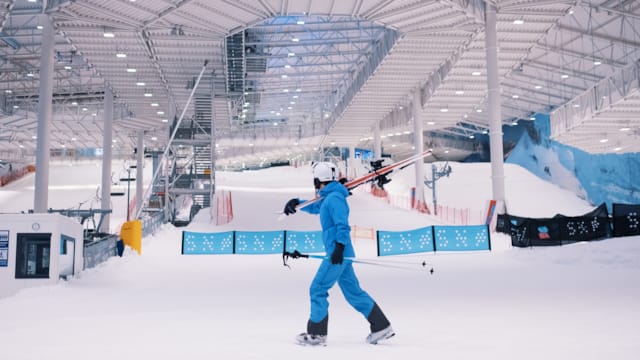 Man wearing blue ski suit carries a pair of skis in the Thon Snø indoor ski arena