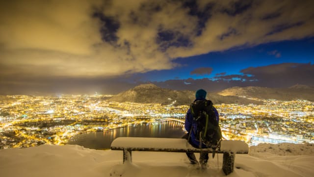 Man sitting on bench looking towards a scenic view of illuminated Bergen city in the night