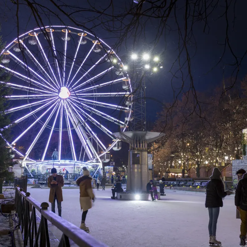 Nattscene med opplyst pariserhjul på vinterfestivalen med folk som går på snødekt sti og festlige høytidslys.