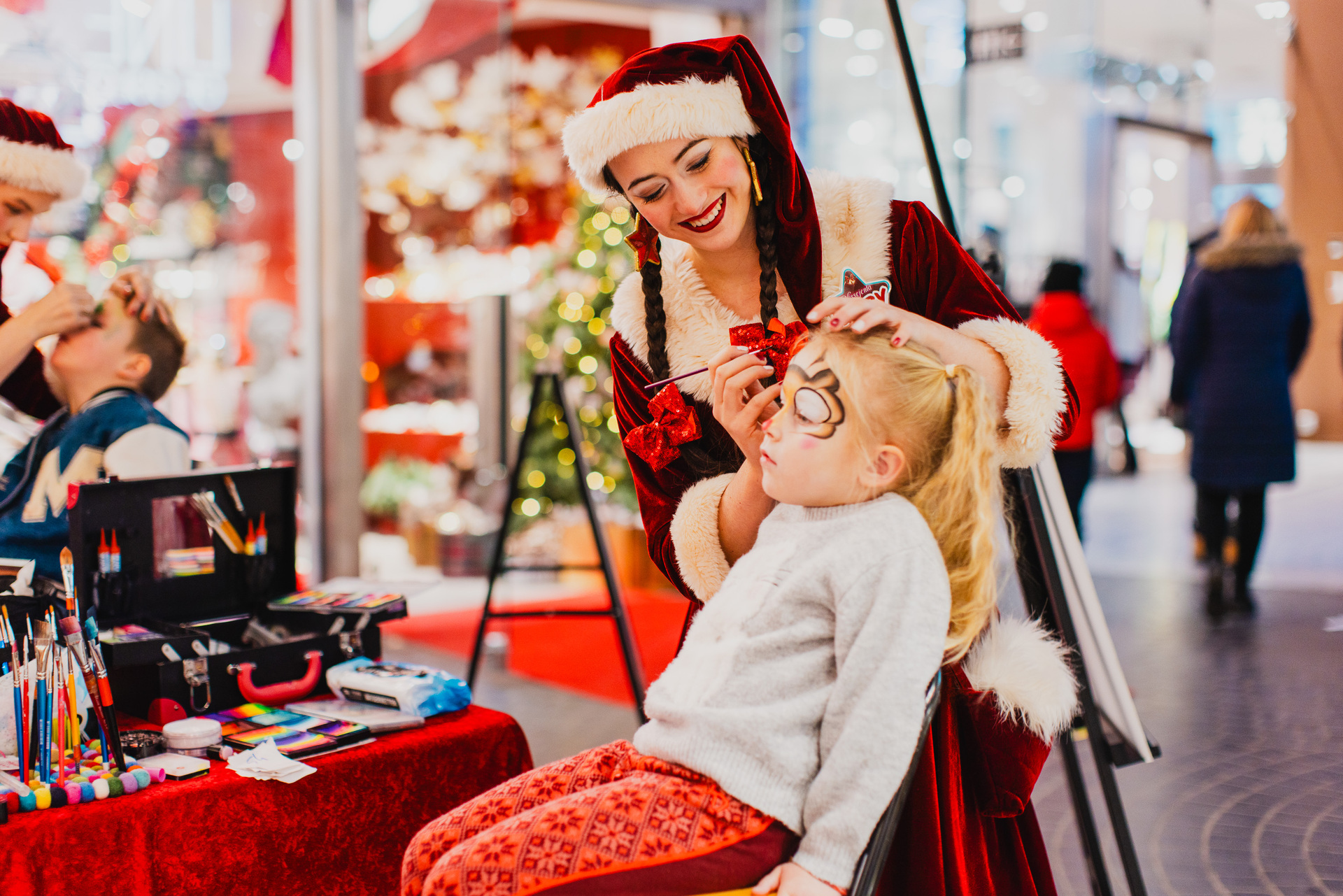 A woman dressed as a Christmas elf doing face painting on a young child at a Christmas market.