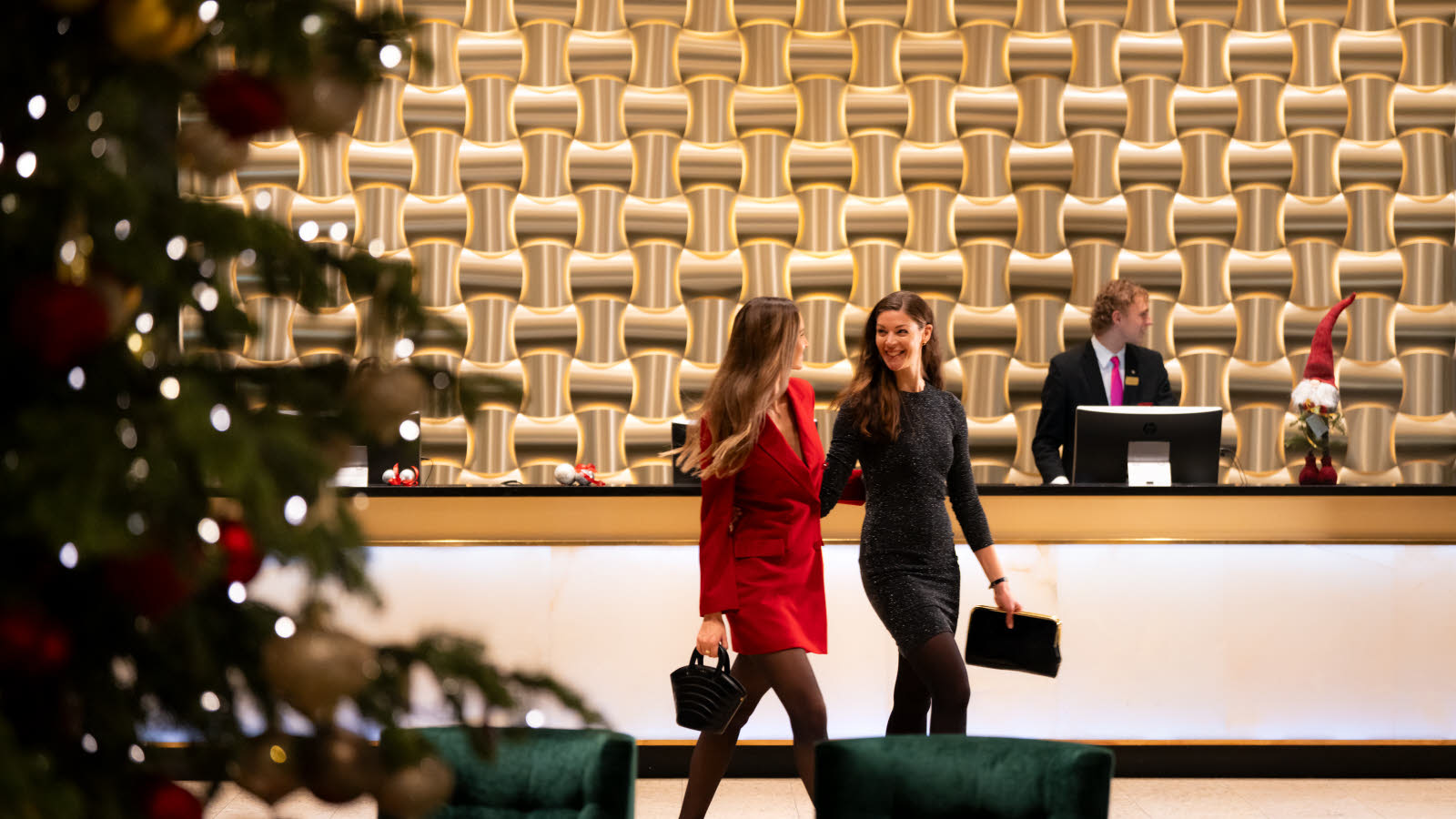 Two well-dressed women walking in front of a hotel receptiondesk decorated for Christmas. They are talking and laughing together.