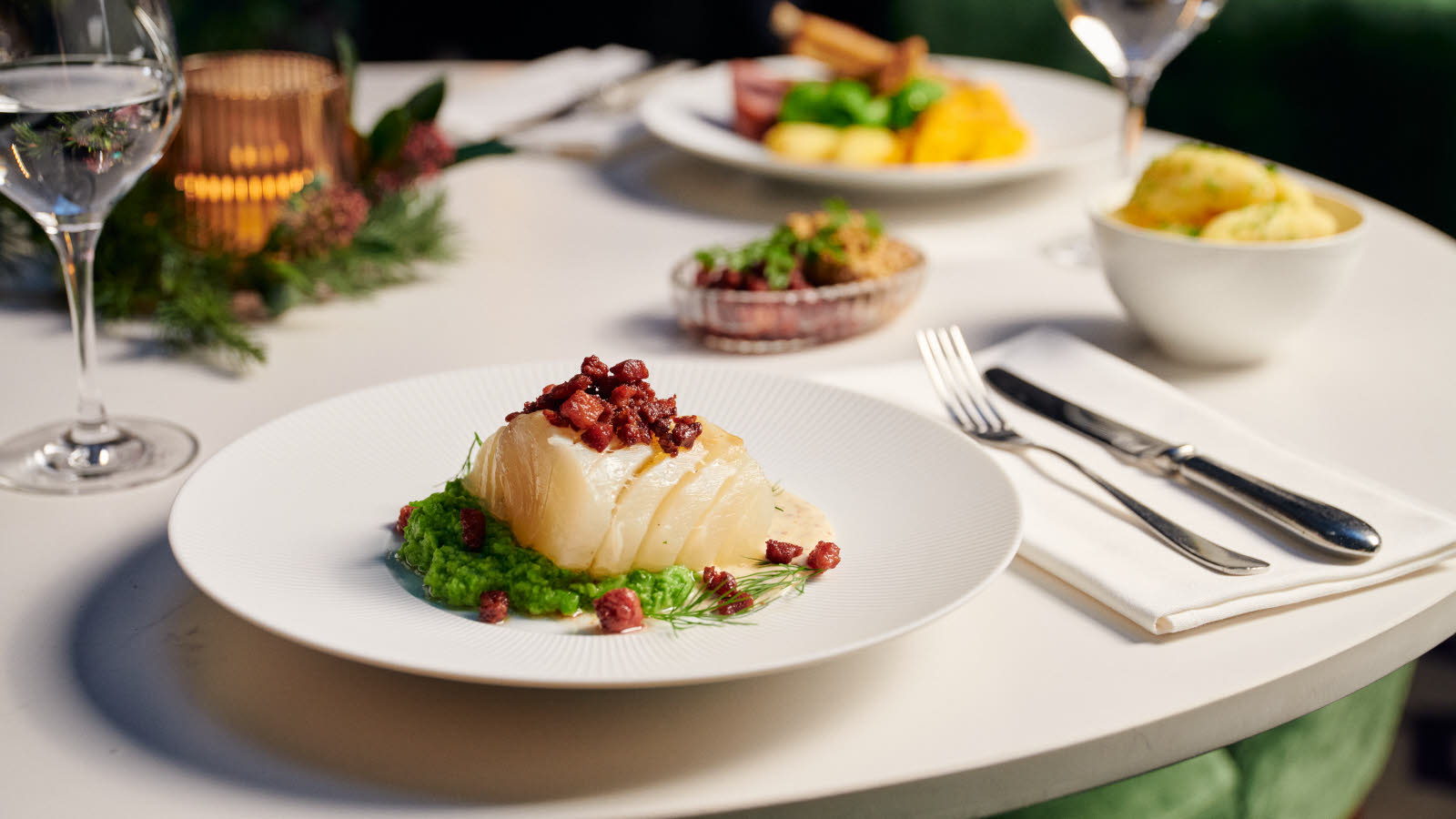 Utsnitt av hvitt bord med tallerken med lutefisk og ertepuré, samt vinglass og bestikk. Flere tallerkener med julemat i bakgrunnen. 
