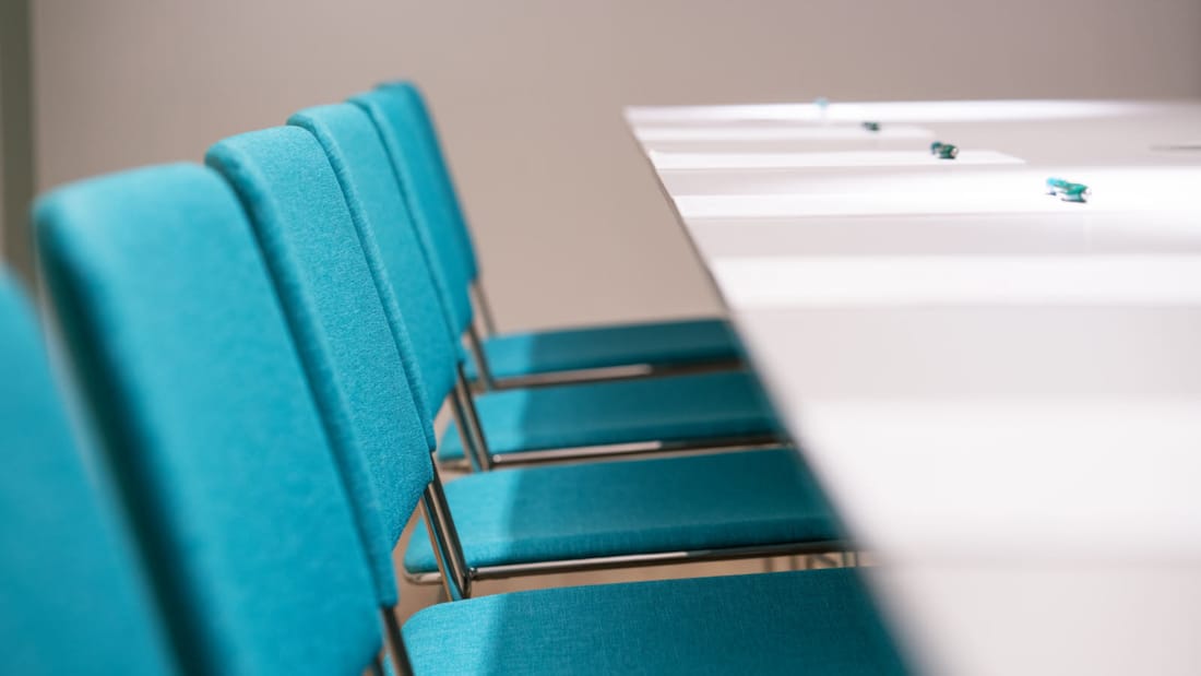Skrivbord och turkosa stolar i ett mötesrum.