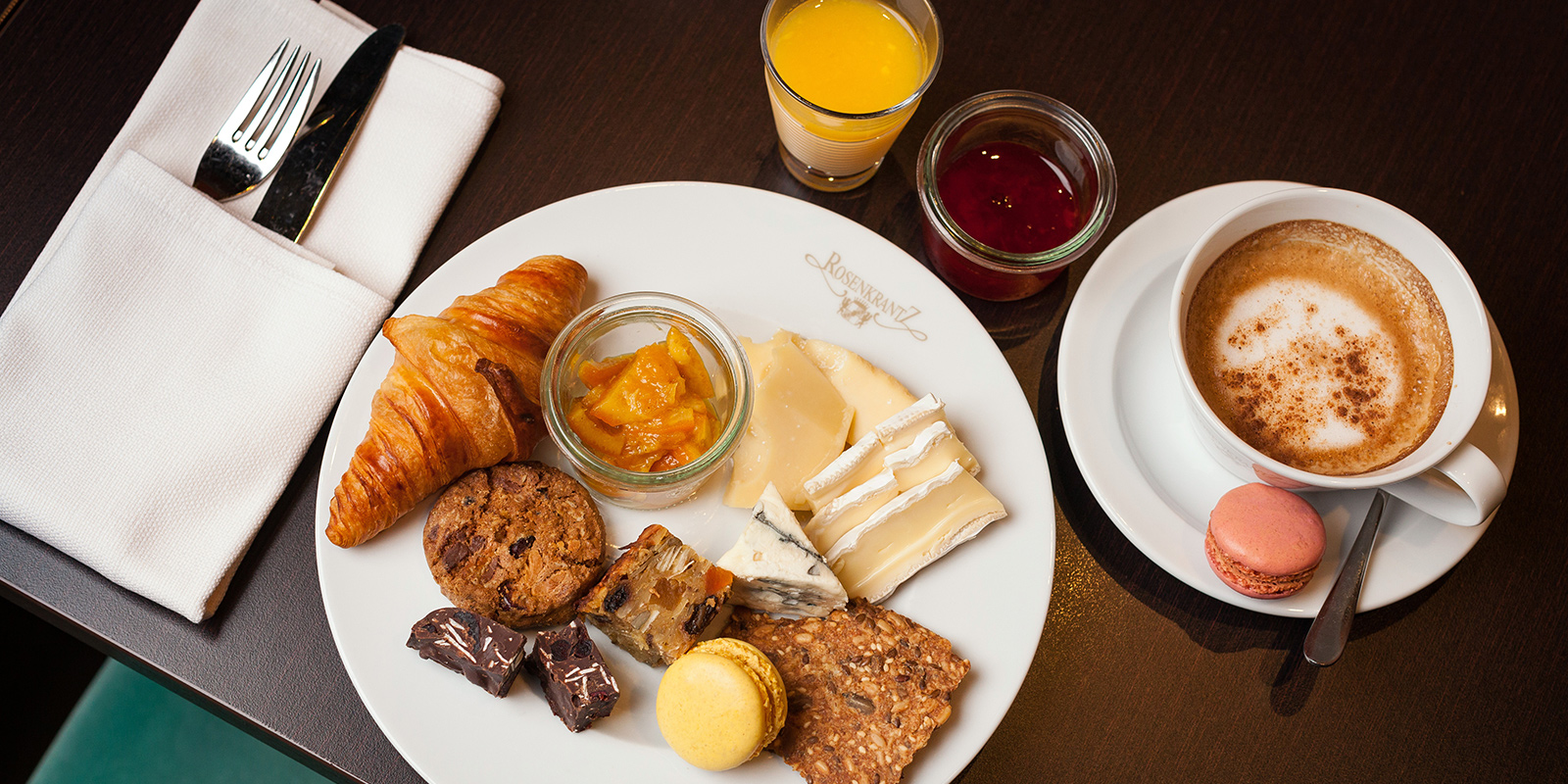 A rich breakfast platter consisting of a croissant, macarons, biscuits and marmelade. Cappuccino and organce juice on the side.