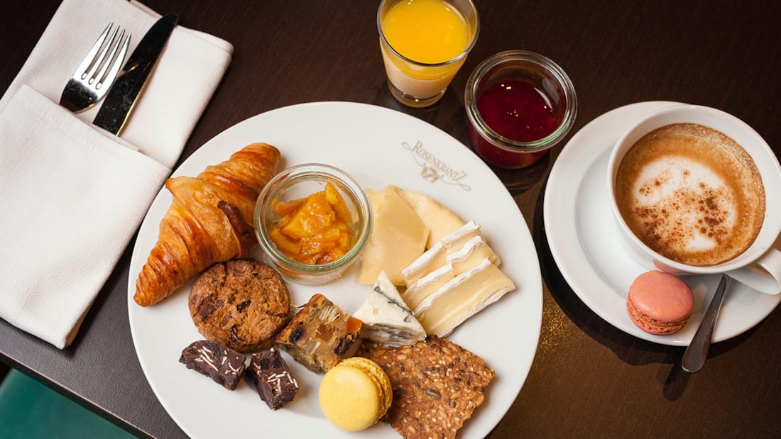 A rich breakfast platter consisting of a croissant, macarons, biscuits and marmelade. Cappuccino and organce juice on the side.