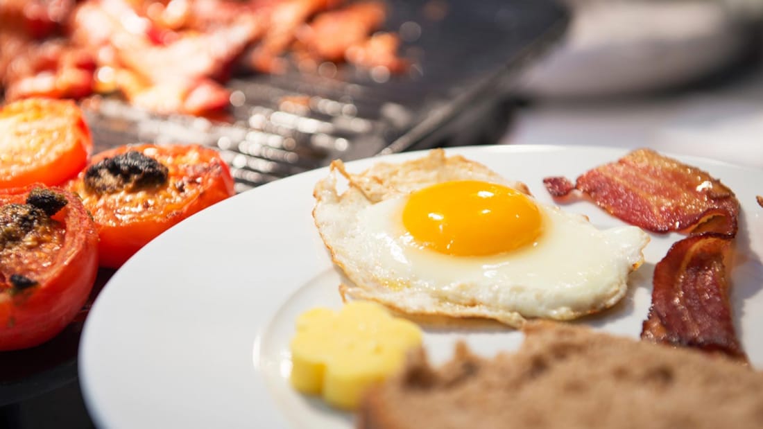 Detail image of a sunny side up fried egg, bacon, a piece of toast and ovenbaked tomatoes topped with pesto.