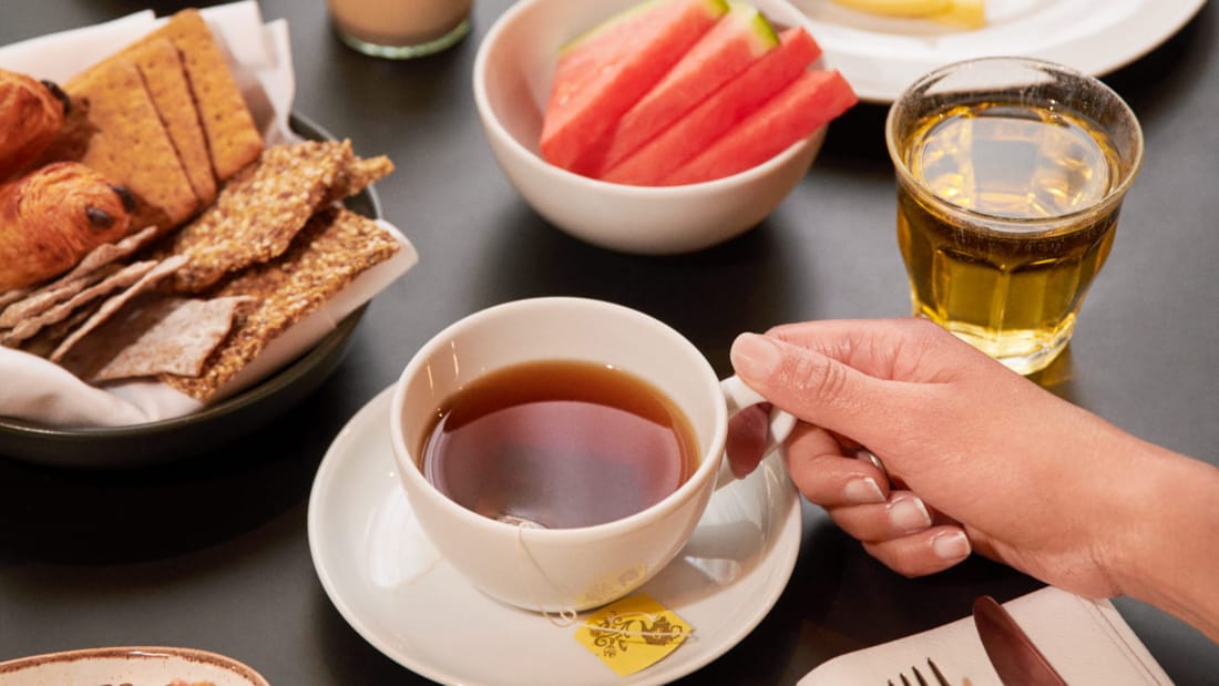 Een hand tilt net een theekop op van een ontbijttafel.