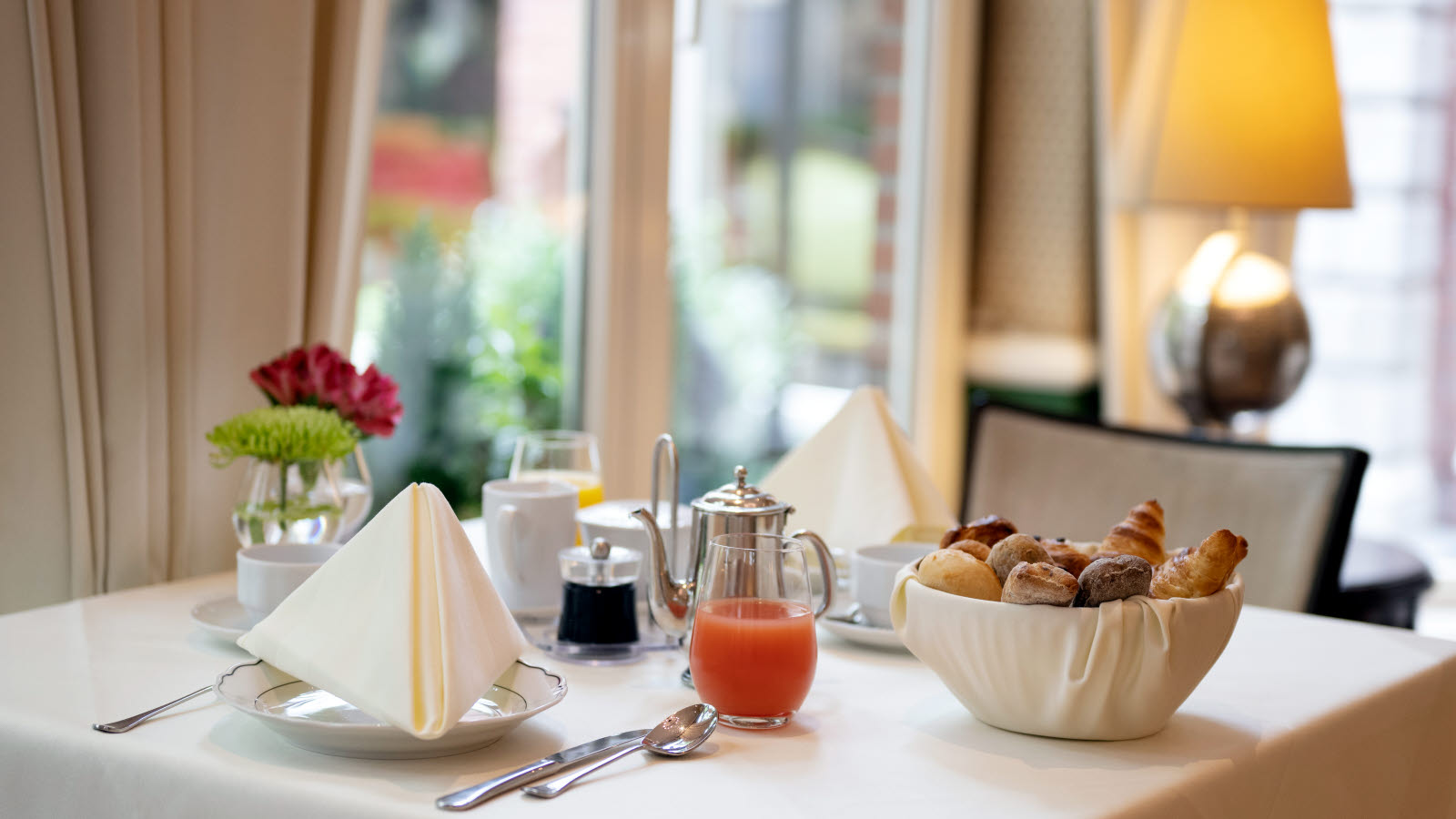 Et bord med et hvitt duk, dekket med serviser, juice og en kurv med rundstykker