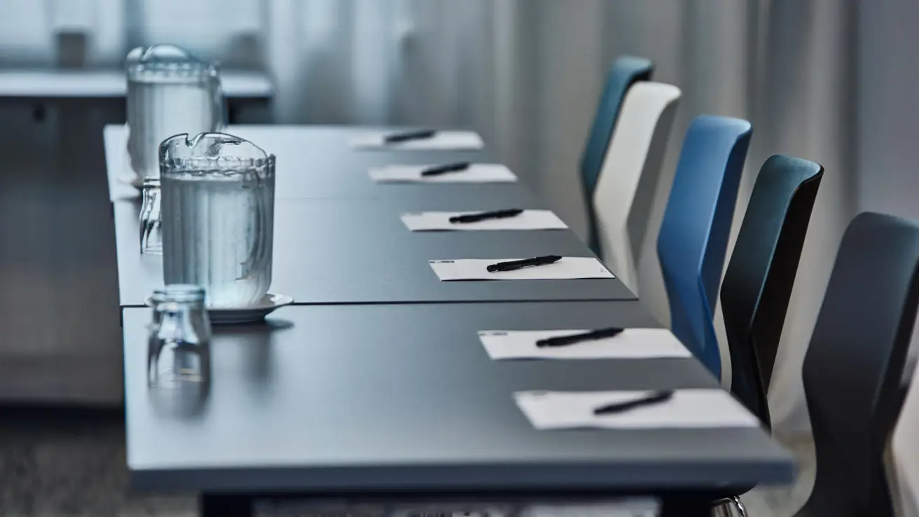 Gros plan sur un bureau et des chaises. La table est recouverte de papeterie, de verres à eau et de carafes à eau.
