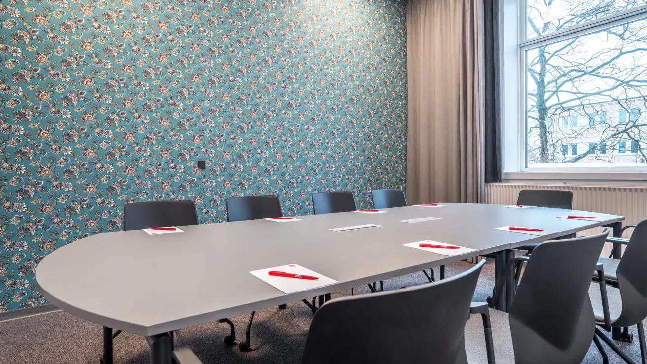 Table avec 9 chaises de bureau sur roulettes tout autour. Papeterie sur la table. Fenêtre et rideaux. Papier peint bleu avec un motif floral en rouge, blanc, vert et jaune.
