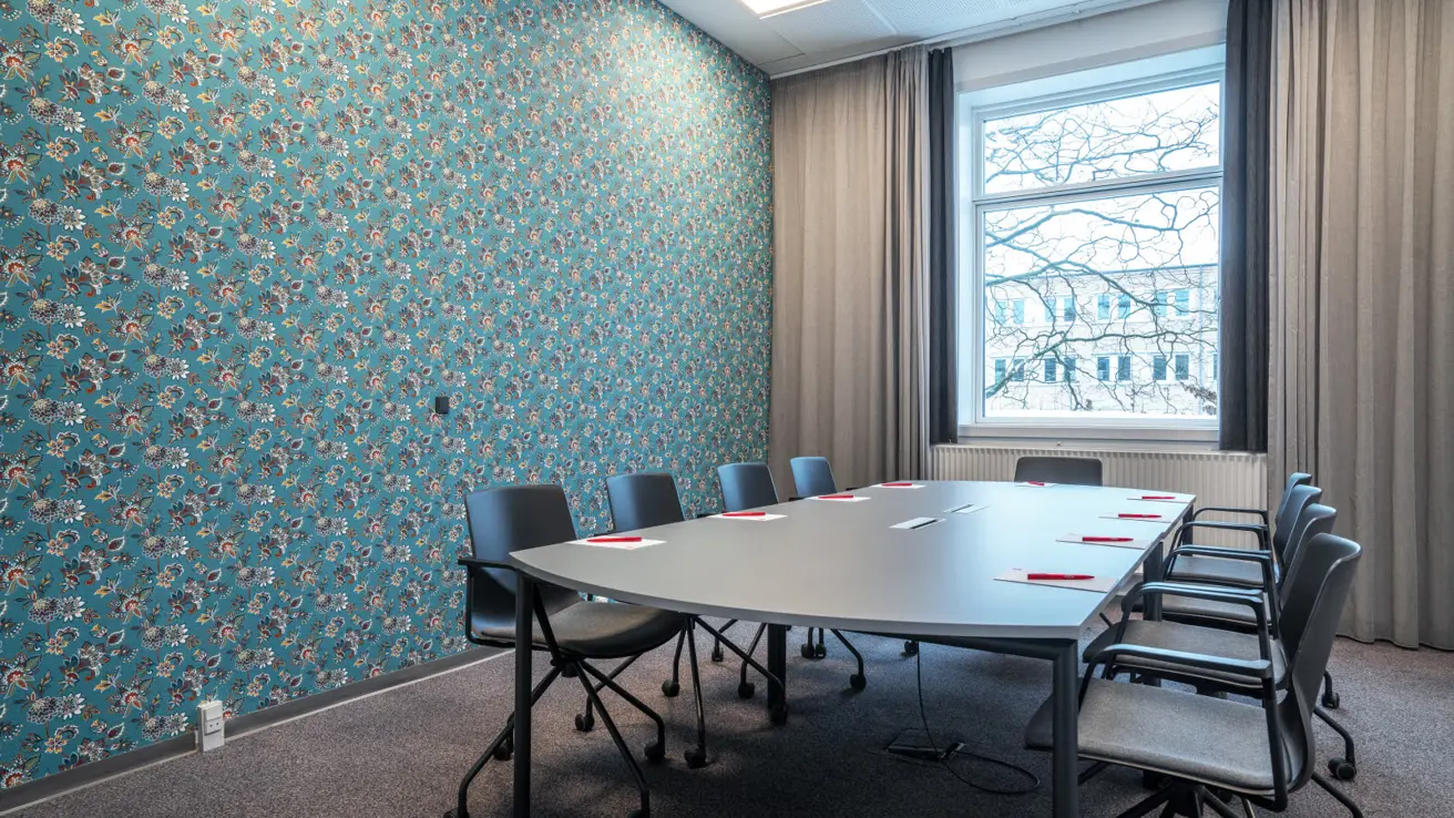 Table avec 9 chaises de bureau sur roulettes tout autour. Papeterie sur la table. Fenêtre et rideaux. Papier peint bleu avec un motif floral en rouge, blanc, vert et jaune.