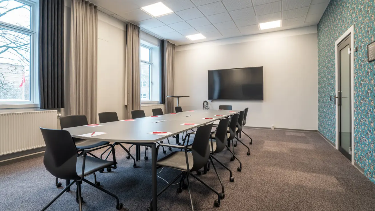 Salle de réunion 8 avec deux fenêtres sur le mur long, écran TV sur le mur court, disposition d’une table de conseil avec douze chaises de bureau sur roulettes tout autour. Mur de papier peint et porte vitrée à droite.
