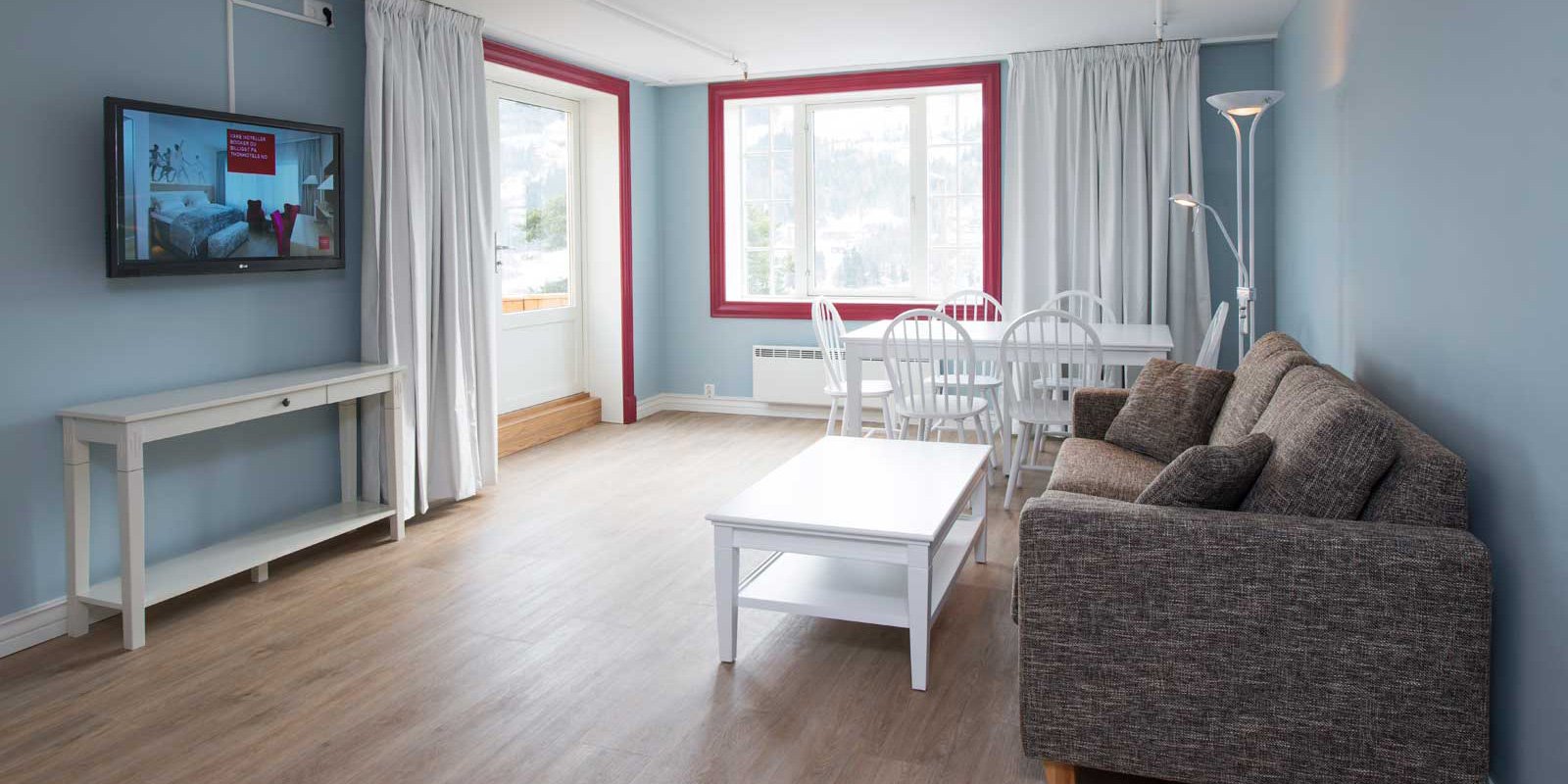 Suite with brown sofa, white coffee table, TV on the wall, two windows with white curtains, blue walls, parquet floor.