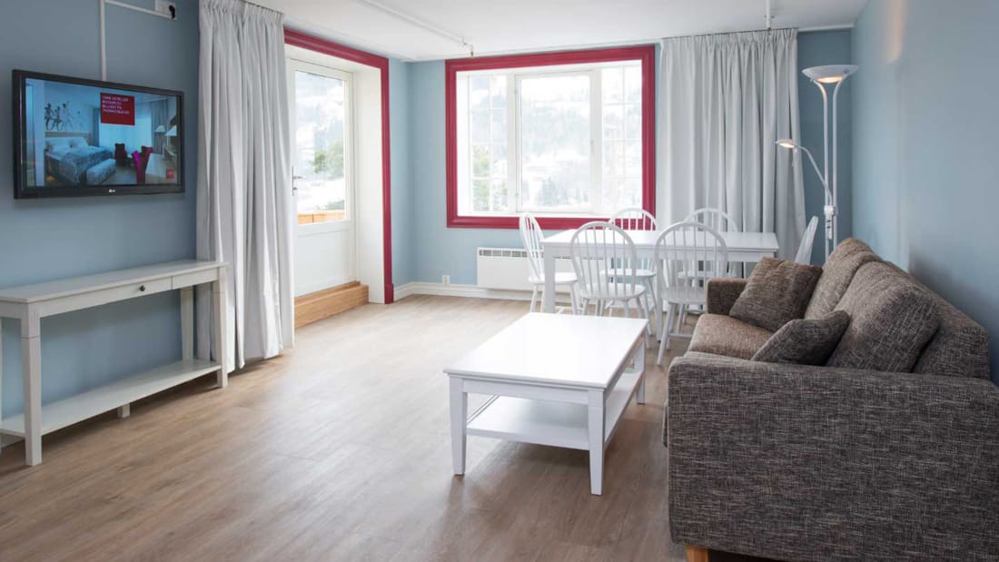 Suite med brun sofa, hvitt salongbord, TV på veggen, to vinduer med hvite gardiner, blå vegger, parkettgulv.