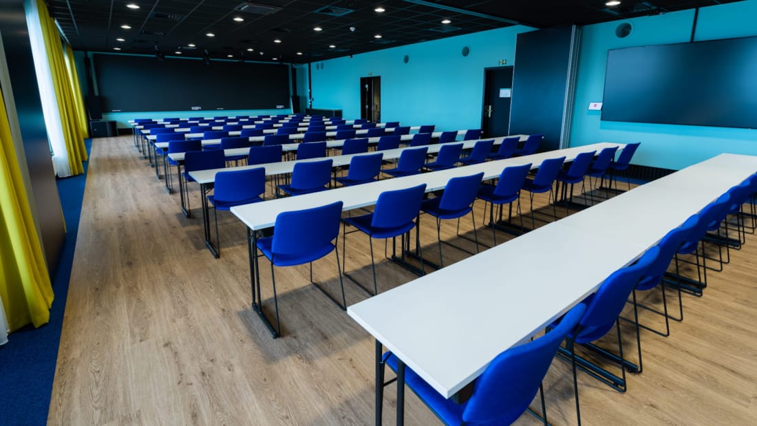 Konferensrum i klassrumslayout med blå stolar, TV-skärm och svart tavla