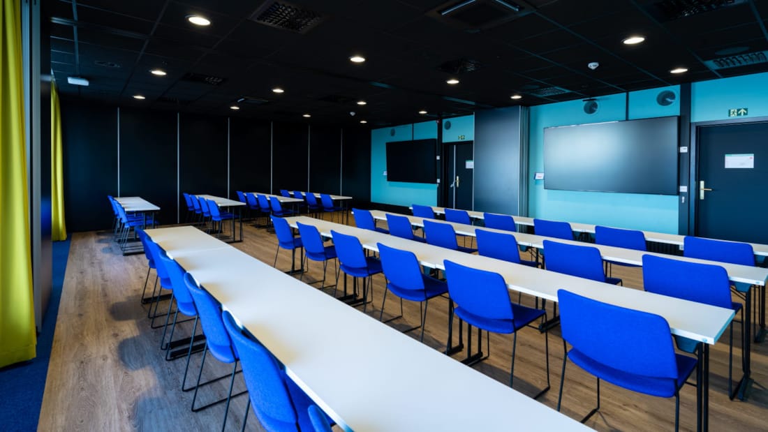 Mødelokale i klasseværelse med blå stole og to tv-skærme