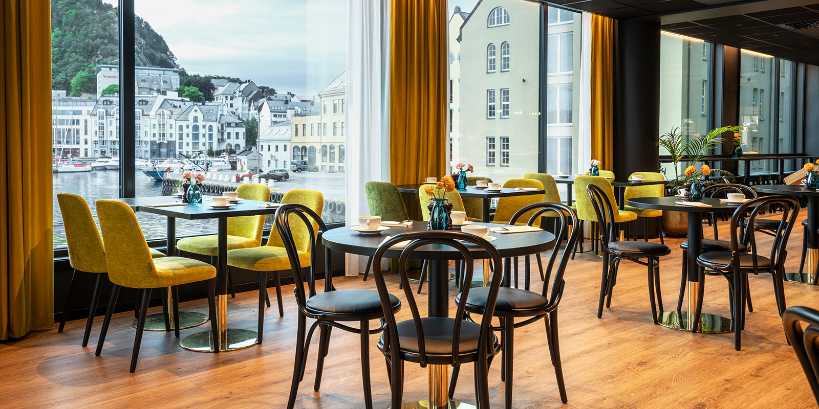 Restaurant med runde bord og stoler foran store vinduer med utsikt over Ålesund by.