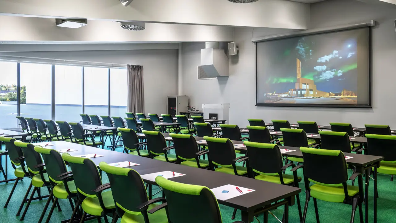 Desks, white wall with projector screen, window with grey curtains, floor-to-ceiling window, green carpet, green chairs.