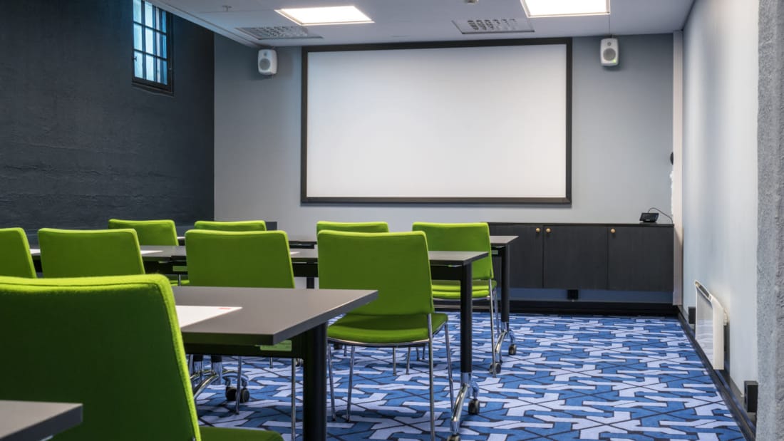 Mødelokale med en række grønne stole og sorte borde foran en stor skærm med højtaler
