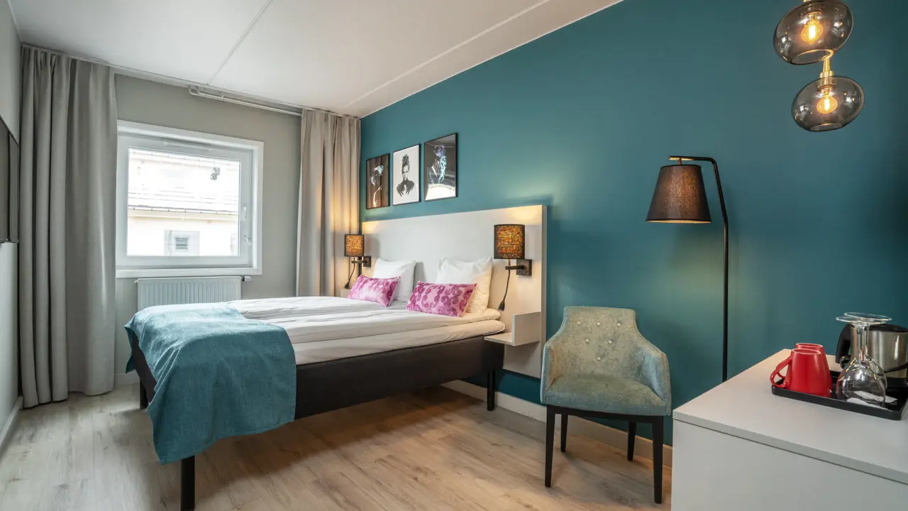 Doppelzimmer mit großem Bett vor einer türkisfarbenen Wand mit Fenster und Schreibtisch.