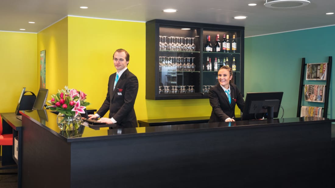 Resepsjon med gule og turkise vegger med to smilende resepsjonister på Thon Hotel Gardermoen