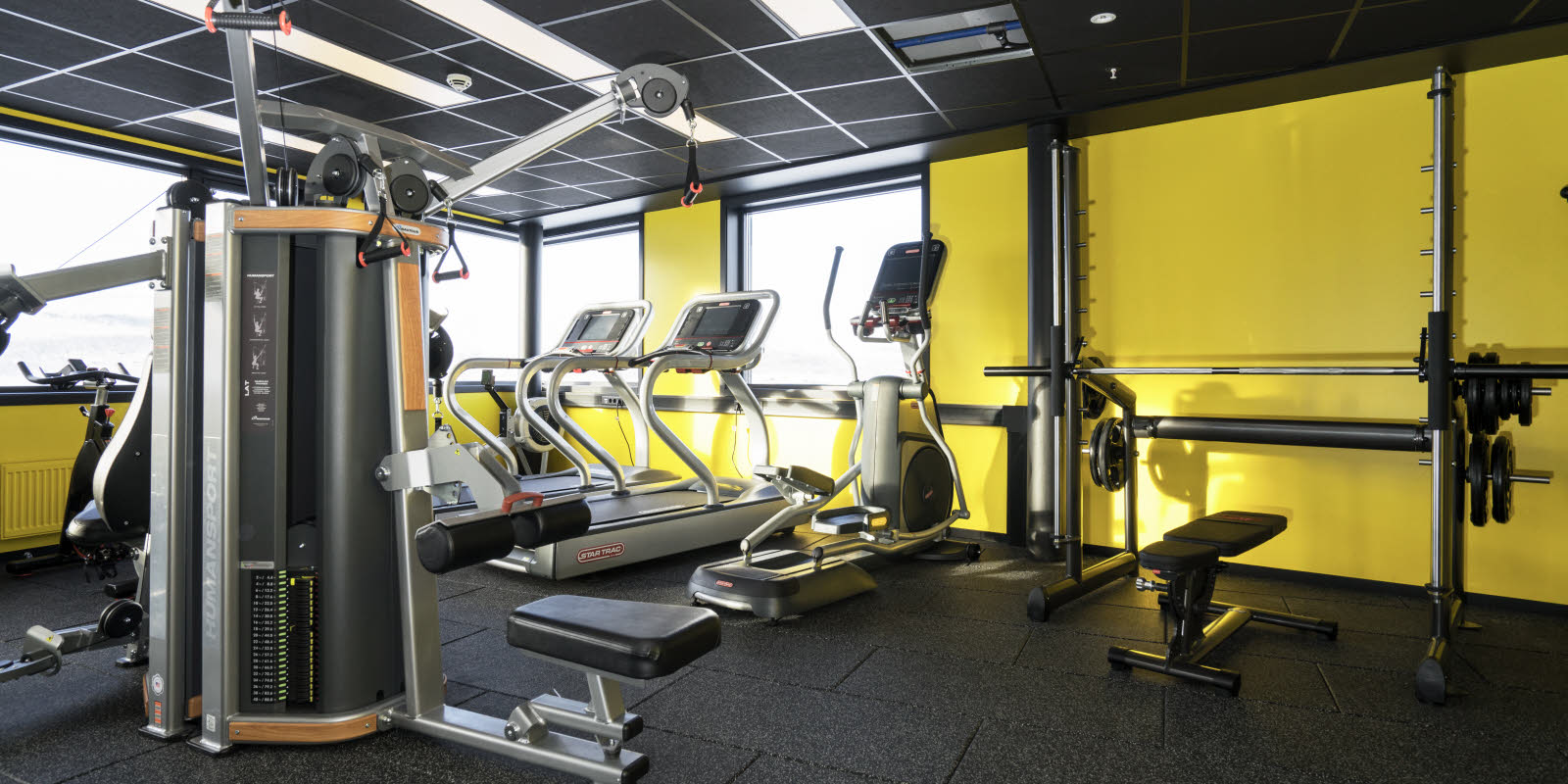 Fitness-rum med forskellige træningsmaskiner, gul væg og udsigt fra vinduet på Thon Hotel Kirkenes ved kajen i Kirkenes centrum