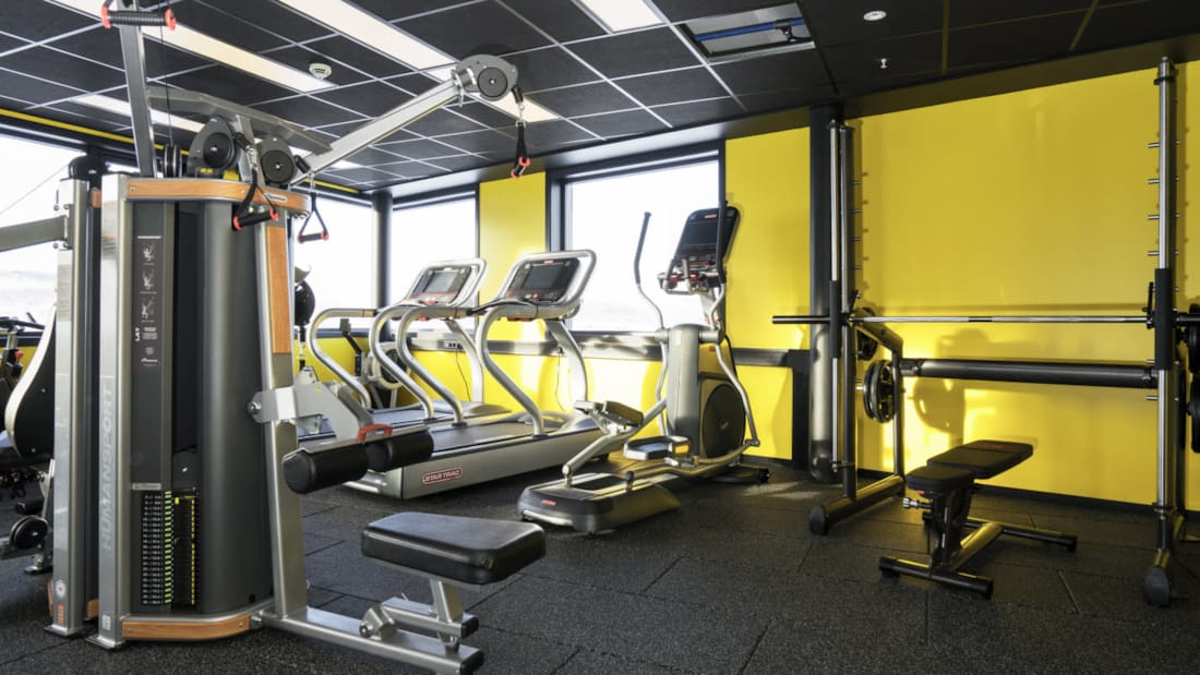 Fitness-rum med forskellige træningsmaskiner, gul væg og udsigt fra vinduet på Thon Hotel Kirkenes ved kajen i Kirkenes centrum
