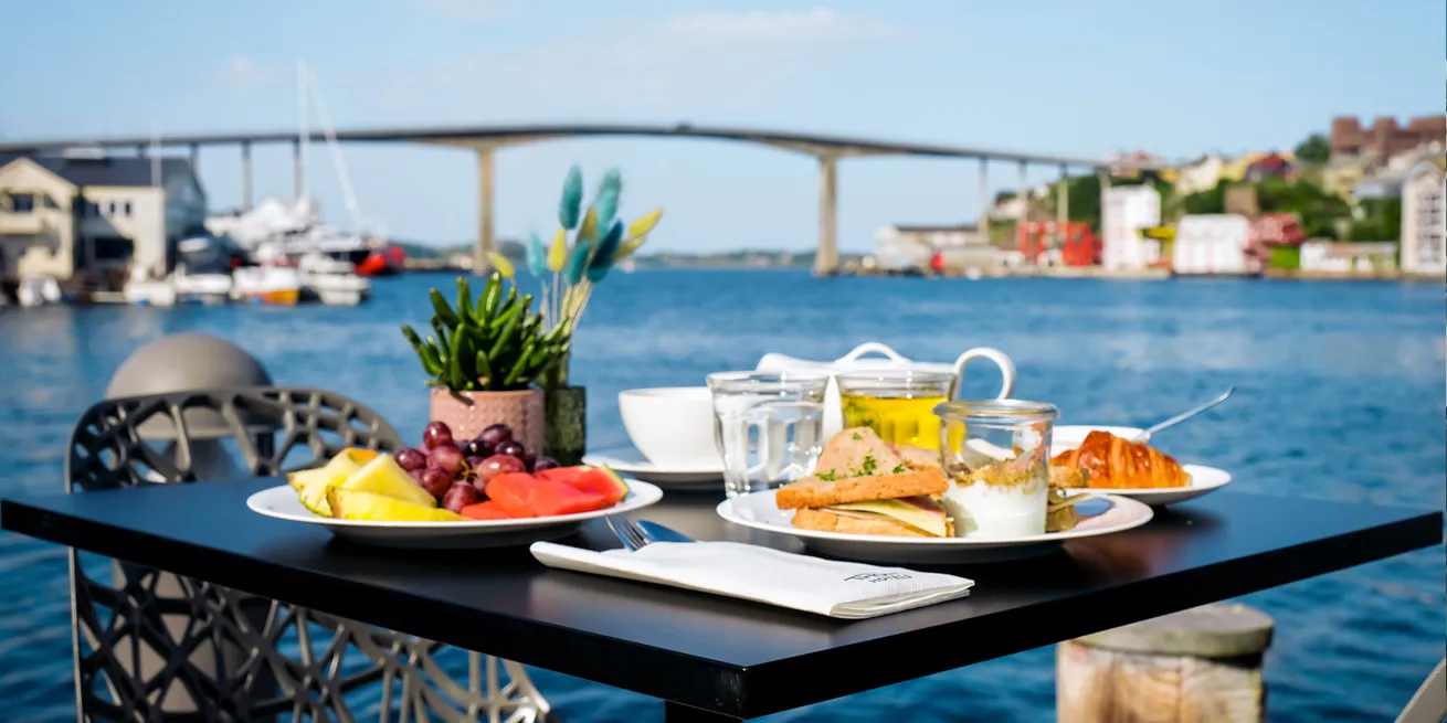 Frokostbord p&aring; en terrasse med frukt, sandwich og kaffe, med utsikt til havet og broen i Molde.