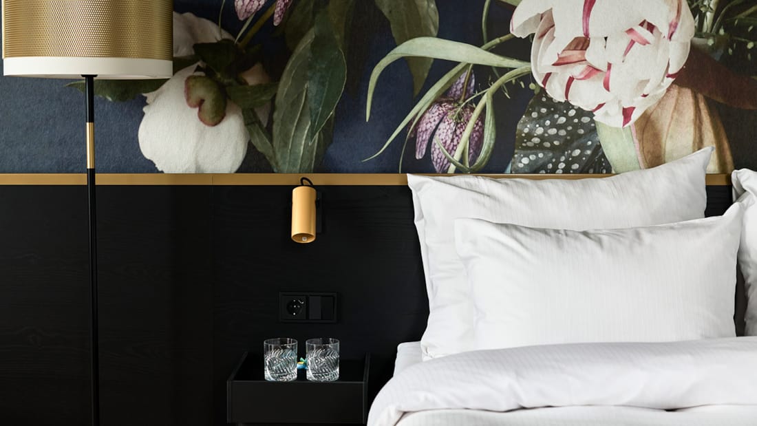 Close-up of made-up bed with bedside table and lamp to the left of the bed. Floral wallpaper behind the bed.