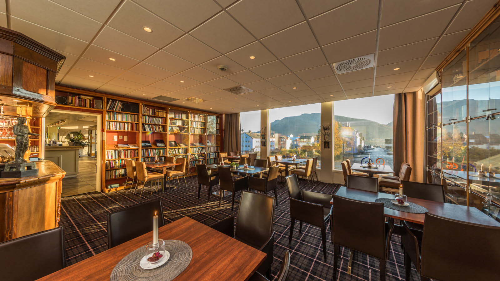 Hotellets bibliotek med flere borde og siddepladser, tæppebelagt gulv og store vinduespartier.