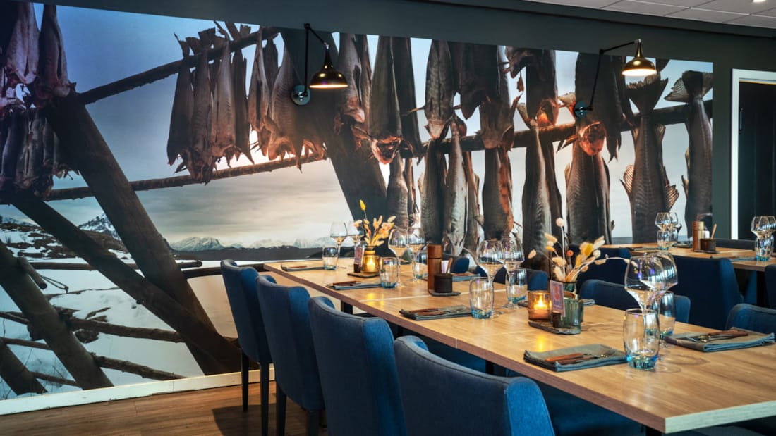 Dukat bord på Sult Grillhouse med glas, bestick, servetter och dekorationer på bordet. På väggen hänger en nästan heltäckande bild av torrfisk upphängd på tork.
