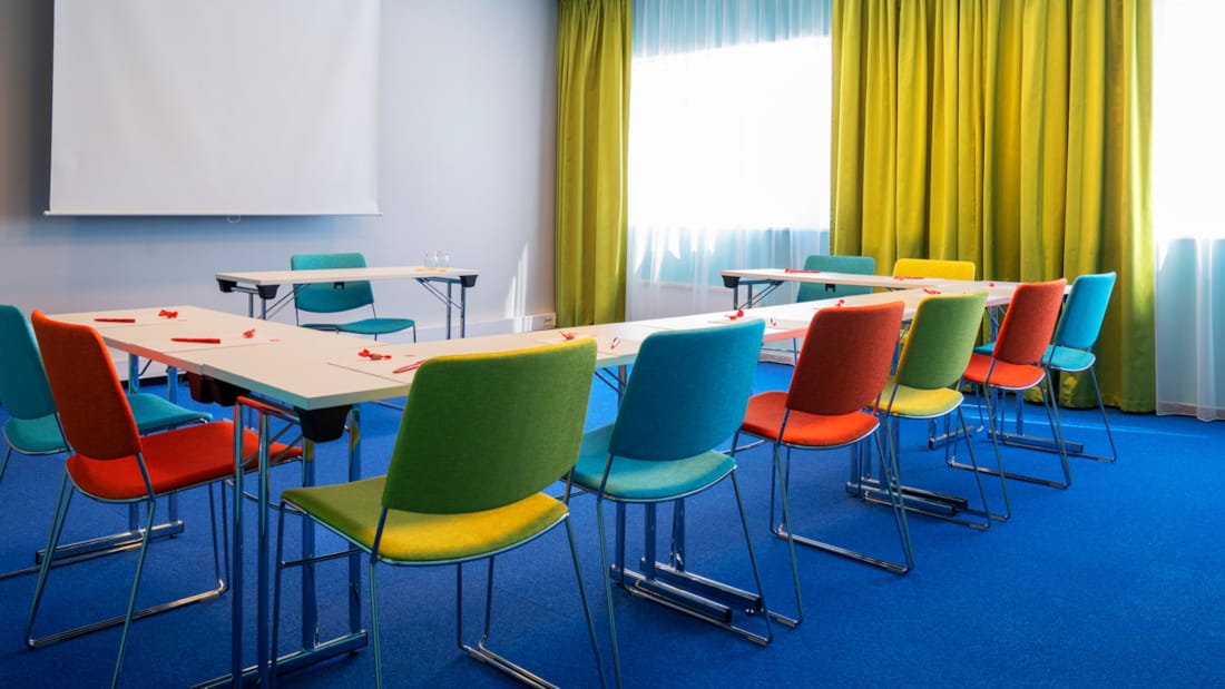Salle de réunion "Øst 6" avec moquette bleue au sol, murs turquoise, rideaux jaune moutarde, écran, projecteur et chaises colorées en forme de fer à cheval au Thon Hotel Triaden à Lørenskog