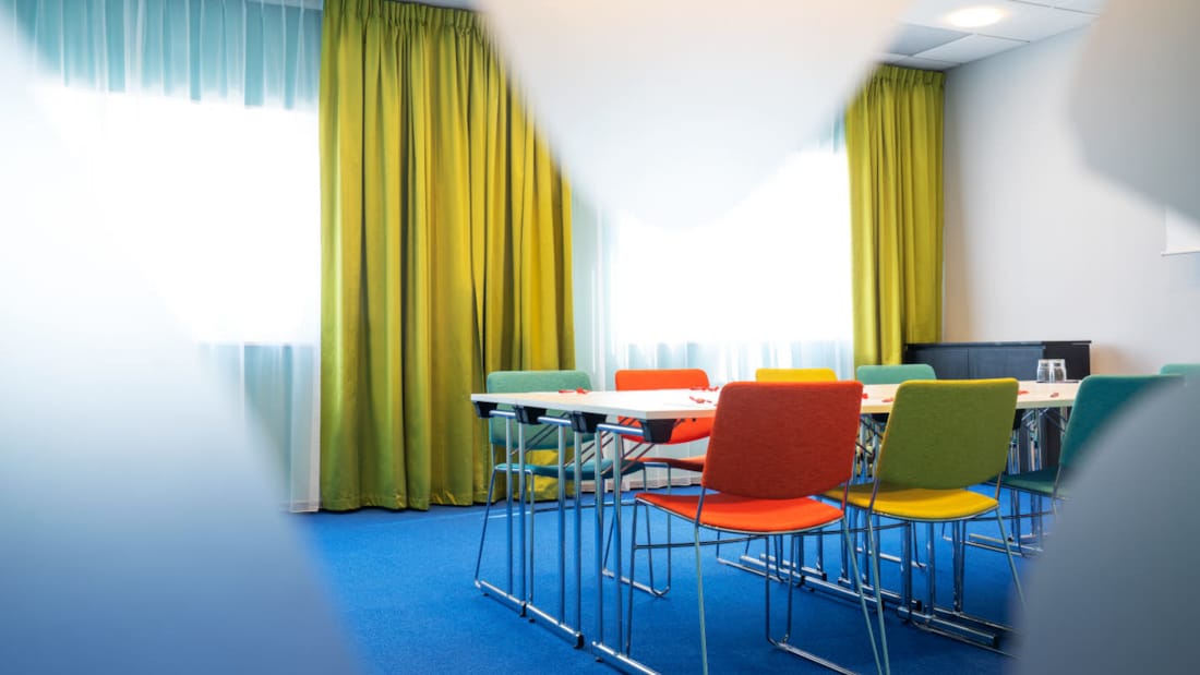 Mötesrum Øst 6 med blå matta på golvet, turkosa väggar, senapsgula gardiner, duk, projektor och färgrika stolar med styrelserumsmöblering på Thon Hotel Triaden i Lørenskog