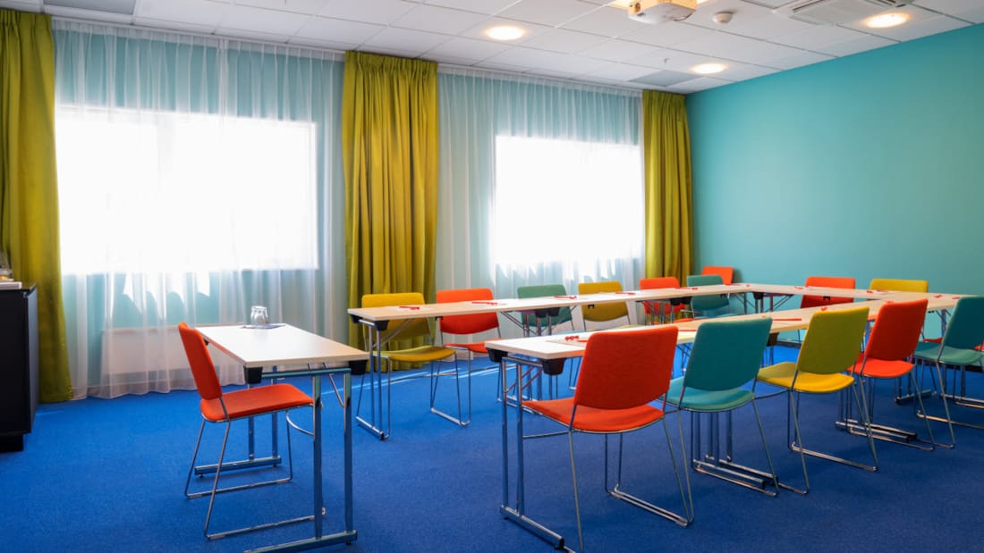 Salle de réunion "Øst 5" avec moquette bleue au sol, murs turquoise, rideaux jaune moutarde, écran, projecteur et chaises colorées en forme de fer à cheval au Thon Hotel Triaden à Lørenskog