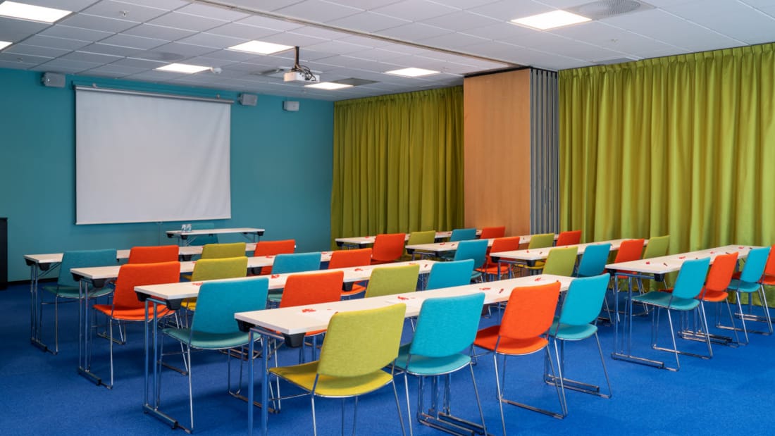 Meeting room "Øst 2" with blue carpet on the floor, turquoise walls, mustard yellow curtains, canvas, projector and colorful chairs in a classroom layout at Thon Hotel Triaden in Lørenskog