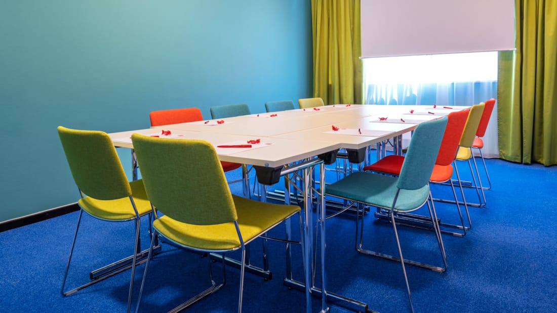 Salle de réunion "Øst 8" avec moquette bleue au sol, murs turquoise, rideaux jaune moutarde, écran, projecteur et chaises colorées dans une disposition de table de conseil au Thon Hotel Triaden à Lørenskog