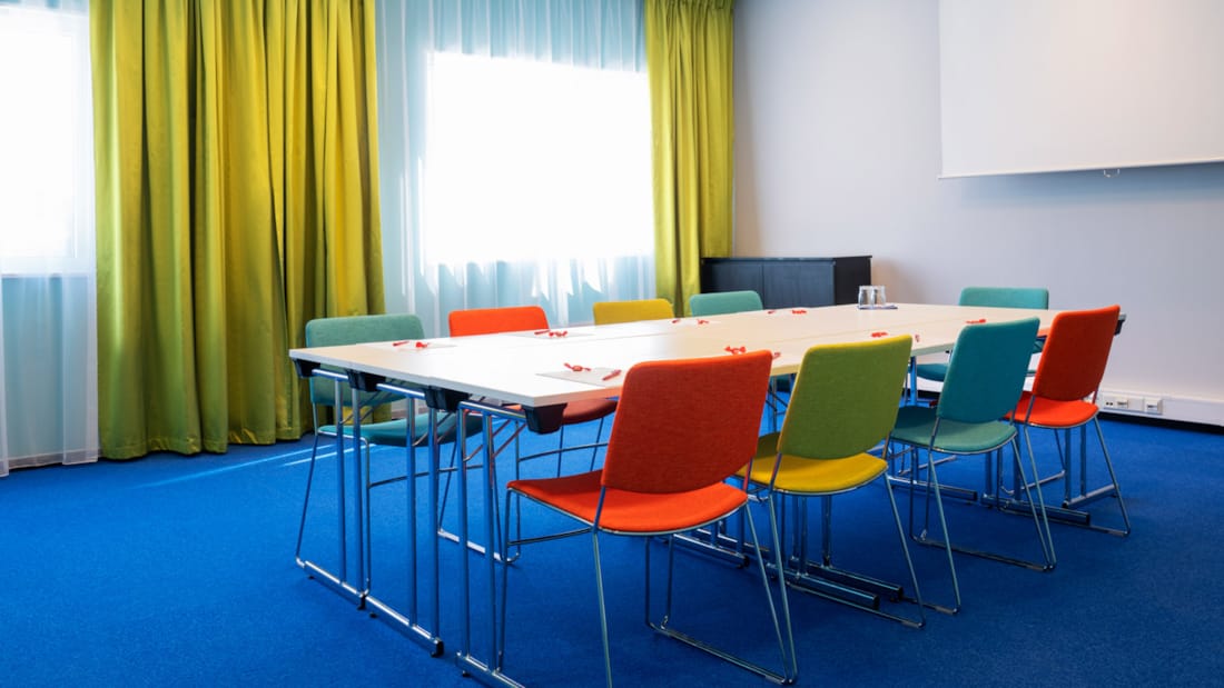 Meeting room "Øst 7" with blue carpet on the floor, turquoise walls, mustard yellow curtains, screen, projector and colorful chairs in a board table layout at Thon Hotel Triaden in Lørenskog
