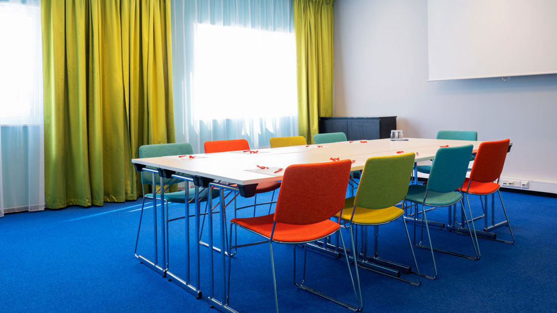Mötesrum Øst 7 med blå matta på golvet, turkosa väggar, senapsgula gardiner, duk, projektor och färgrika stolar med styrelserumsmöblering på Thon Hotel Triaden i Lørenskog