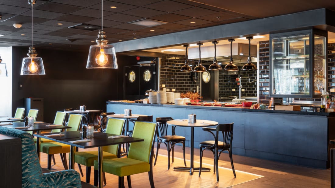 Restaurang med öppet kök i bakgrunden med industrilampor samt bord och stolar i förgrunden på Thon Hotel Triaden i Lørenskog