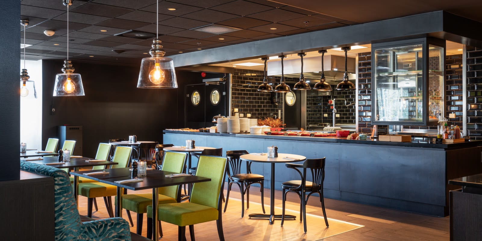 Restaurang med öppet kök i bakgrunden med industrilampor samt bord och stolar i förgrunden på Thon Hotel Triaden i Lørenskog