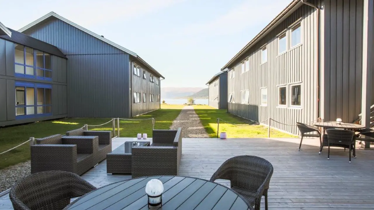 Utsnitt av bord og stoler på en platting, to bygninger i bakgrunnen med utsikt til fjorden mellom seg.