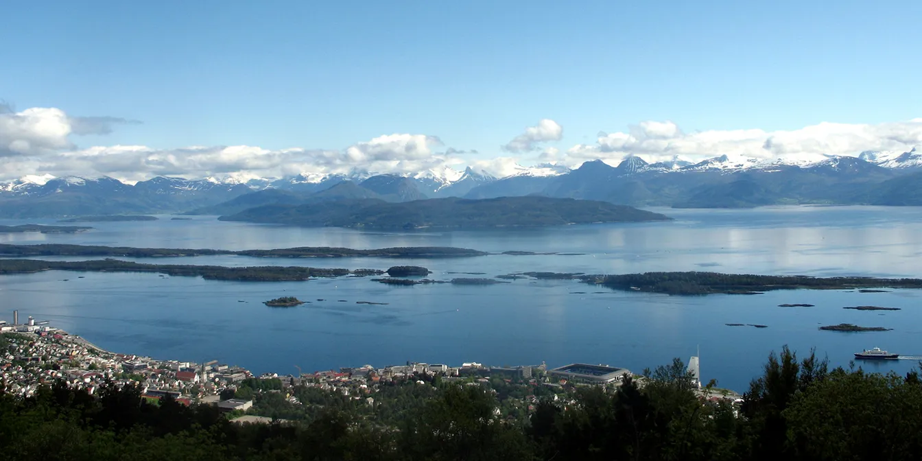 Molde seen from above with majestic mountains on the horizon, white clouds in the sky and blue sea with buildings in front.