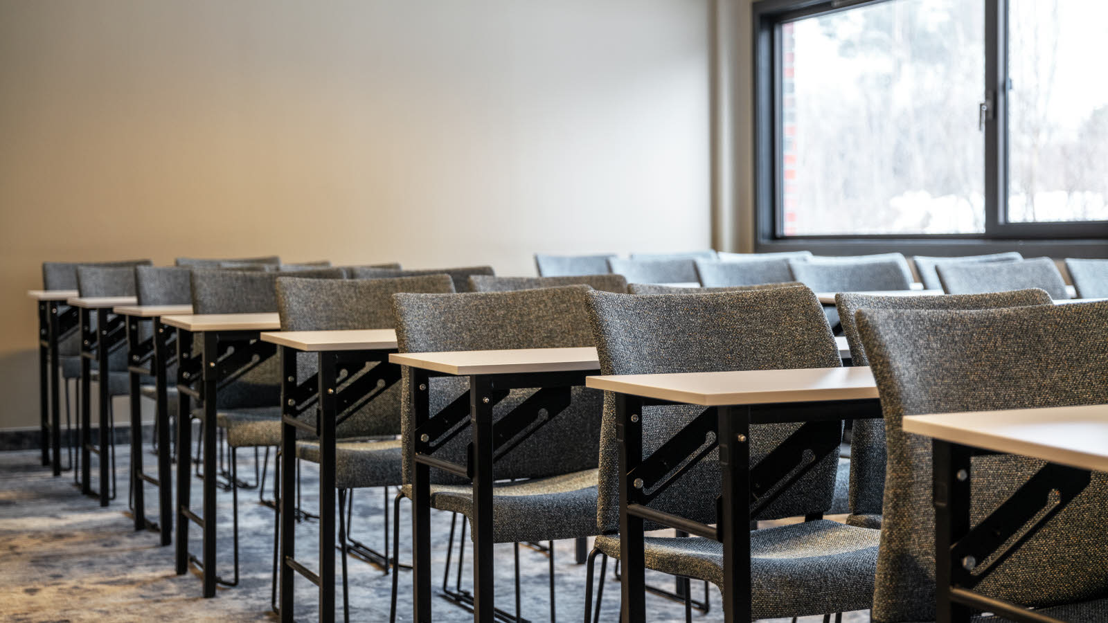 Udsnit af klasseværelsesopsætning i mødelokale. Grå stole, gulvtæppe og store vinduer i den ene side.