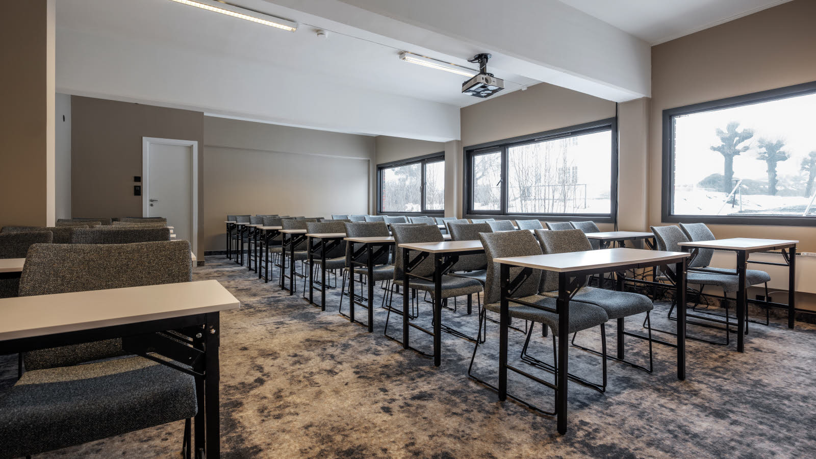 Mødelokale til 42 personer i klasseværelsesopsætning, loftshængt projektor, gulvtæppe, udgang bagerst i lokalet, vinduer på langsiden.