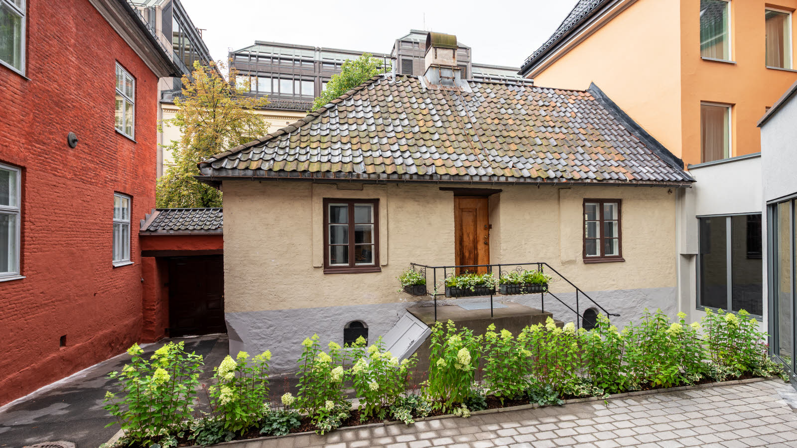 Bilde av et gammelt lite hus som er lokalisert mellom større og mer moderne bygninger.