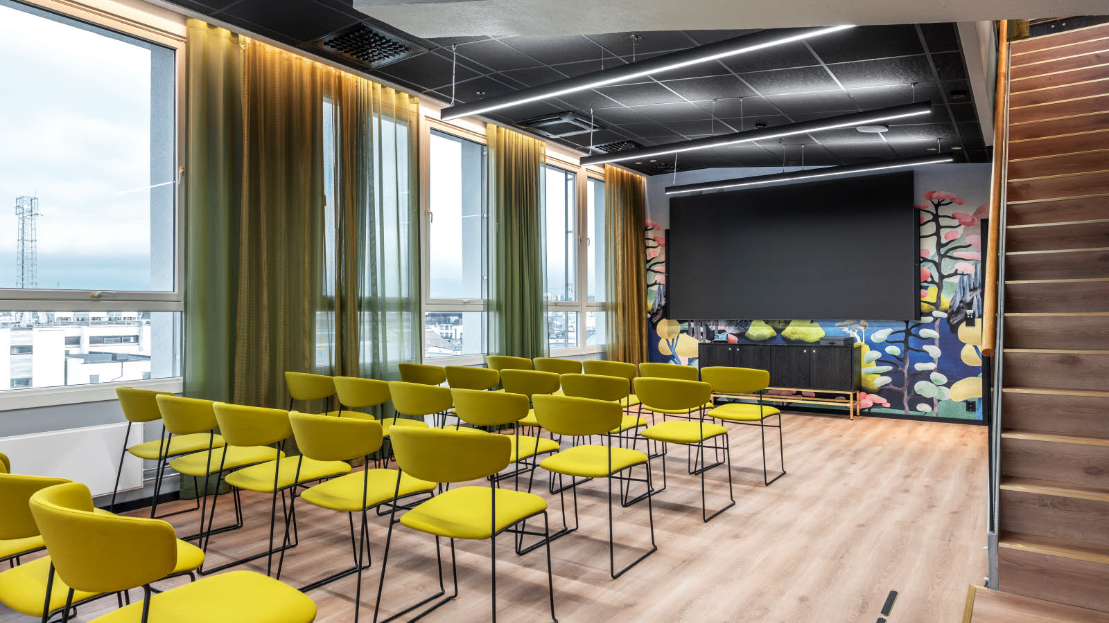 Møteromoppsett med grønne stoler på rekke og rad. Store vinduer langs den ene veggen med utsikt.