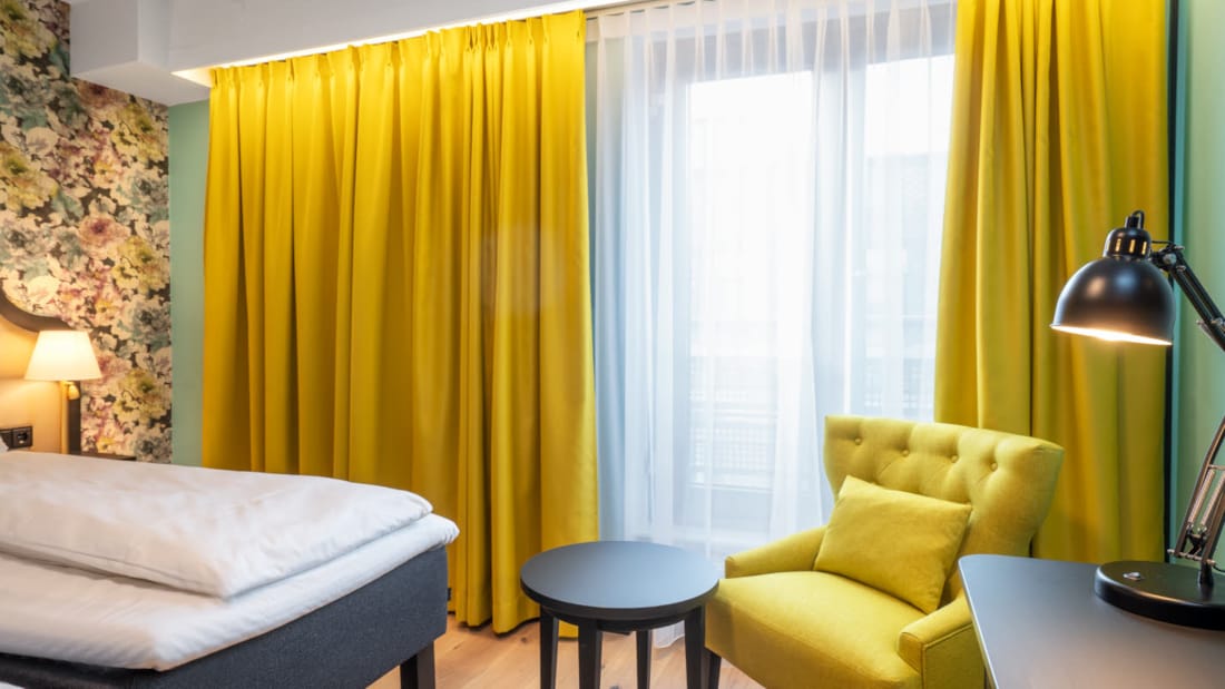 To enkeltsenge, blomstret tapet bag sengene, gul stol, arbejdsbord og store vinduer med store gule gardiner i twin room på Thon Hotel Cecil i Oslo centrum